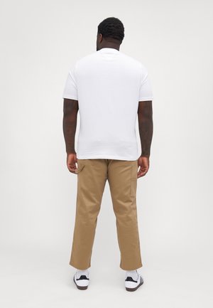 Hombre con tatuajes que lleva una camiseta blanca, pantalones beige con bolsillos laterales y zapatillas blancas, de pie de espaldas contra un fondo liso.