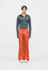 Maglietta grigia a maniche lunghe con collo a mezza zip, abbinata a pantaloni cargo larghi arancioni e scarpe color crema. Design minimalista e vestibilità comoda.