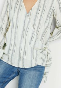 Top ligero de crema con rayas verticales negras, escote en V, corte holgado y detalle atado en el lateral. Combinado con jeans de mezclilla azules.