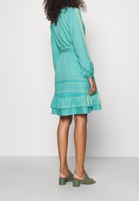 Vestido turquesa con estampado, con un dobladillo con volantes, mangas largas y un cinturón. Combinado con mules de tacón cuadrado verdes.
