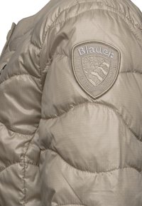 Beige pufferjacka med ett quiltat mönster, som har en texturerad tyg och en broderad logotyp på axeln.