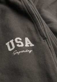 Sudadera con cremallera gris que presenta texto bordado en blanco "USA" y "Superdry" en una fuente casual, fabricada con un tejido suave y con un acabado texturizado.