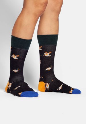 Chaussettes noires avec un revers côtelé vert, ornées de motifs de canards colorés ; les renforts orange au talon et les pointes bleues ajoutent un contraste.