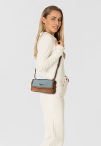 Borsa marrone e blu con motivo geometrico, forma rettangolare, tracolla e dettaglio logo. La modella la indossa con un outfit chiaro.