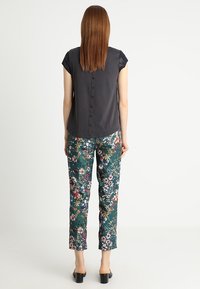 Blusa corta de manga corta en gris oscuro con detalles de botones en la parte posterior, combinada con pantalones verdes con estampado floral, que presentan un ajuste slim y zapatos planos.