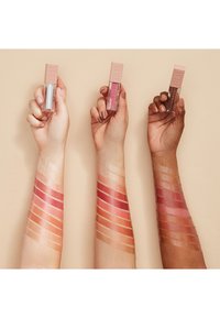 Three hands hold clear lip gloss tubes with light pink caps. Swatches of various shades, from nude to deep pink, are visible on their skin.