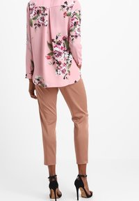 Blouse longue à manches longues rose à motif floral, coupe décontractée, associée à un pantalon beige fuselé et des sandales noires à talons hauts.