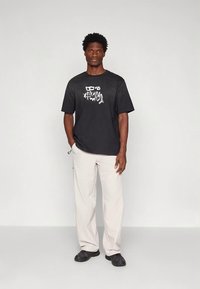 Černé tričko s krátkým rukávem s bílou grafikou, spárované s béžovými kalhotami a černými teniskami, stojící na neutrálním pozadí.