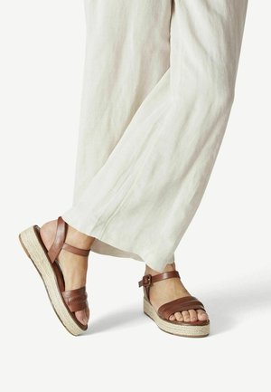 Braune Leder-Wedge-Sandalen mit einer geflochtenen Jutesohle, ausgestattet mit einem Schnallenriemen und strukturierten Akzenten. Tragen Sie sie mit hellfarbigen, fließenden Hosen.