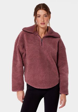 Kobieta nosząca bluzę polarową w kolorze mauve z zamkiem do połowy i prążkowanym kołnierzem, zestawioną z czarnymi spodniami, stojąca na jednolitym tle.