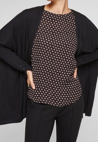 Black cardigan over a patterned top featuring a dark background with small, leaf-like shapes in white and peach. Slim black pants.