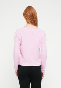 Ροζ πουλόβερ με μακριά μανίκια, στρογγυλή λαιμόκοψη και ριμπ μανσέτες. Το υλικό φαίνεται να είναι μαλακό πλεκτό; το τελείωμα φτάνει στη μέση. Φορεμένο με μαύρο παντελόνι.