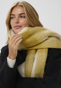Sciarpa rigata gialla e bianca realizzata in materiale morbido e peloso, appoggiata intorno al collo, abbinata a un cappotto scuro testurizzato e a un maglione chiaro.