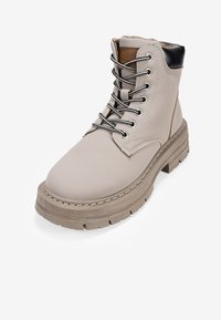 Bottines beige avec un laçage, accent de talon noir et semelle en caoutchouc crantée. Comprend des coutures blanches le long de la semelle et une tige texturée.
