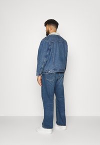 Džinsinė striukė su lengva fliso apykakle, klasikinio mėlyno skalbimo, ir plačios formos džinsai. Modelis dėvi baltus sportbačius, matomas iš nugaros.