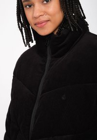 Schwarze Cordjacke mit hohem Kragen, ausgestattet mit einem vorderen Reißverschluss und abgestepptem Design, präsentiert an einem Modell mit geflochtenem Haar.