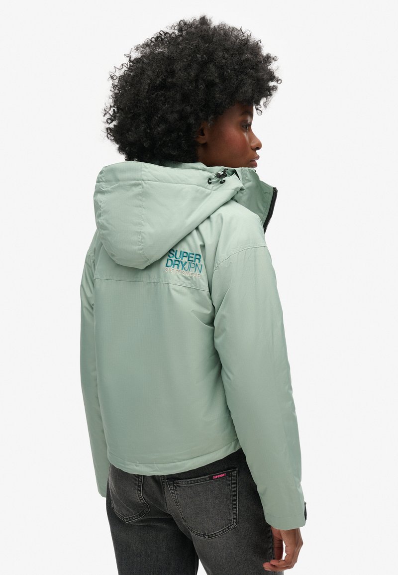 Mintgroene pufferjack met een capuchon, voorzien van een logo op de rug, een gladde textuur en een getailleerd ontwerp. Gekenmerkt door het dragen van donkere spijkerbroeken.