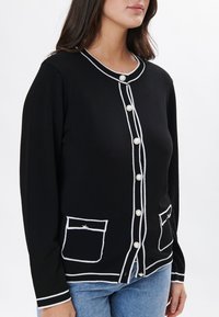 Cárdigan negro con ribetes blancos, presenta botones parecidos a perlas, dos bolsillos frontales y un escote redondo. Material de punto suave.