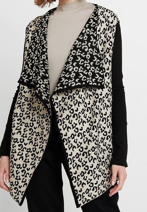 Kobieta ubrana w bezrękawnikowy, beżowo-czarny kardigan w panterkę z otwartym przodem, na czarnej bluzce z długim rękawem i beżowym golfie.