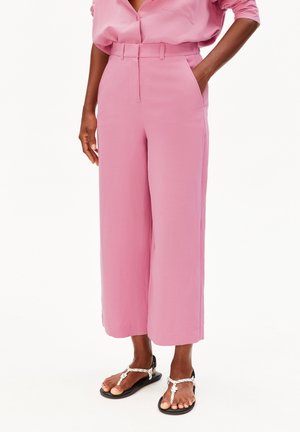 Persoon draagt hoge, wijde roze broek met zijzakken en witte sandaal met gevlochten bandjes, staat met één hand in de zak.