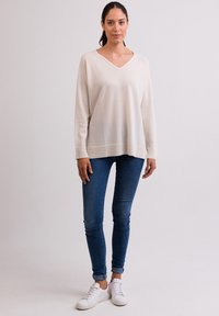 Maglione a maniche lunghe, di colore crema e dal taglio morbido con scollatura a V e orlo a coste, abbinato a jeans blu attillati e sneakers bianche.