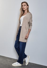 Beige Strickjacke mit offenem Front und Rippbündchen, getragen über einem weißen Oberteil und kombiniert mit blauen weiten Jeans und weißen Sneakern.