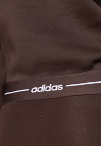 Το καφέ αθλητικό ύφασμα έχει μια ομαλή υφή με το λευκό λογότυπο της Adidas στη μέση, που τονίζεται από μια λεπτή λευκή ρίγα.