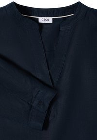 Chemise bleu marine à manches longues avec encolure fendue et poignets boutonnés, avec une étiquette blanche "CECIL" au col.