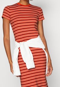 Červené a bílé pruhované šaty s kulatým výstřihem. Mají krátké rukávy a v pase jsou přivázané bílé tričko pro akcent.