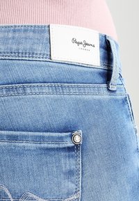 Ljusblå denimjeans med en vit läderetikett som visar "Pepe Jeans London." Funktioner inkluderar sömnad på bakfickorna och metallknappdetalj.