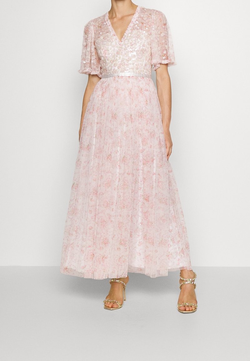 Femme portant une robe longue jusqu'au sol, rose pâle à fleurs, avec un corsage à sequins, des manches courtes bouffantes et des sandales dorées à talons aiguilles ornées de strass.
