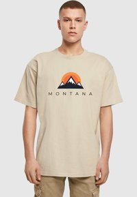 Béžové bavlněné tričko s krátkým rukávem, s grafickým designem hor a slunce v oranžové a černé barvě. Text zní "MONTANA."