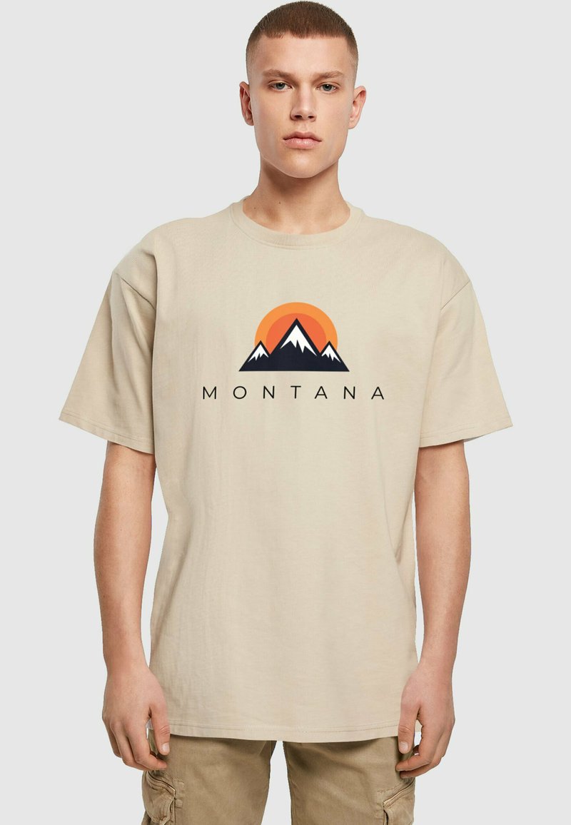 Bežové bavlnené tričko s krátkymi rukávmi, na ktorom je grafický dizajn hôr a slnka v oranžovej a čiernej farbe. Text znie "MONTANA."