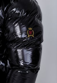 Chaqueta acolchada negra brillante con diseño de acolchado, con un parche de logo visible en dorado, rojo y negro en la manga.