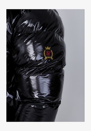 Mustan läikepinnaga tikkpooder jacket, millel on ruuduline disain ja varrukal nähtav kuldne, punane ja musta logopatch.