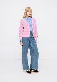 Cardigã rosa de malha, camisa riscada azul, calças de ganga largas e ténis pretos com detalhes em branco. Modelo em pé com as mãos nos bolsos.