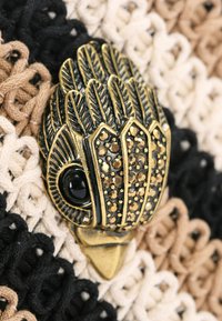 Metalowa broszka w kształcie ptaka, z misternymi detalami piór i czarnym kamieniem szlachetnym w oku, na teksturowanym neutralnym tle z plecionki.