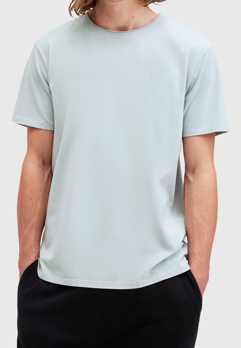 Homme portant un t-shirt gris clair à manches courtes et col rond, les mains dans les poches d’un pantalon noir, cadré au niveau du cou et des hanches.