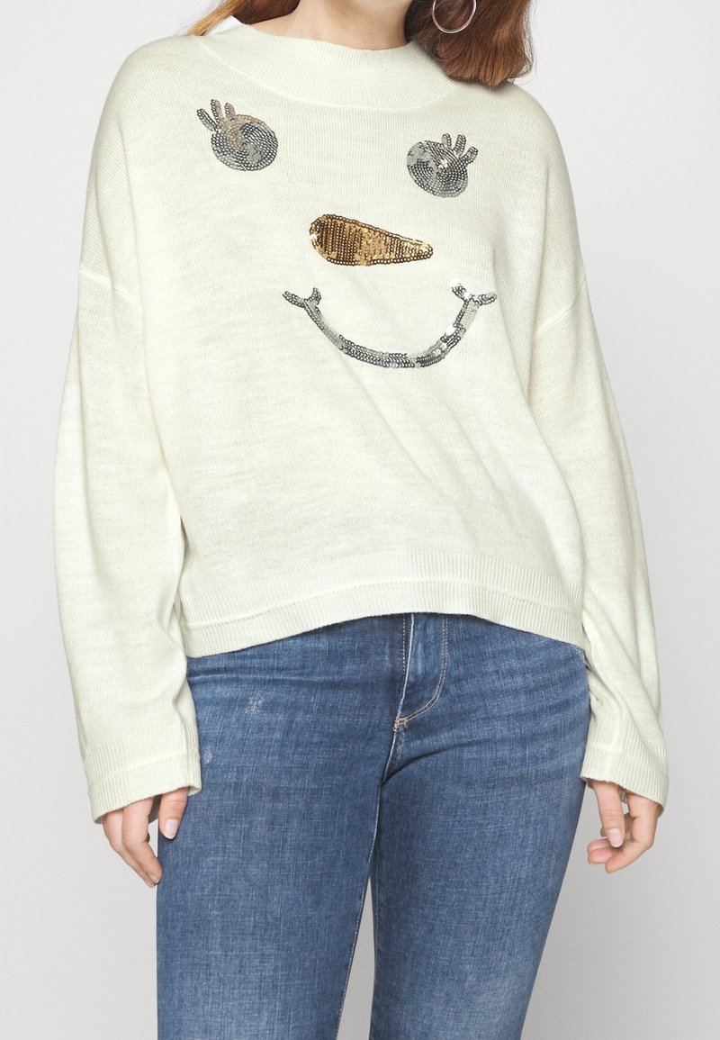 Kobieta w kremowym swetrze z cekinowym wzorem bałwana i niebieskich dżinsach, ukazana od ramion do bioder na białym tle.