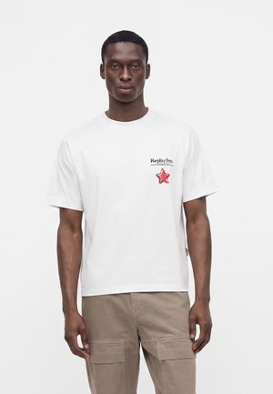 Mann trägt weißes kurzärmeliges T-Shirt mit Replay Inc-Logo und rotem Stern auf der Brust, kombiniert mit beigen Cargohosen, steht vor weißem Hintergrund.