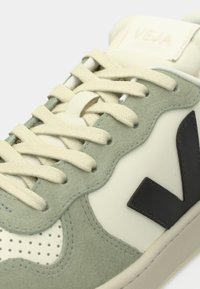 Zapato con parte superior de ante verde claro y cuero blanco roto, con cordones beige y un prominente logo "V" negro en el lateral.