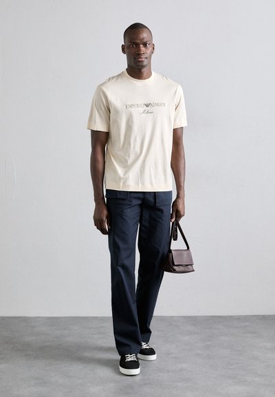 Beige Baumwoll-T-Shirt mit "Emporio Armani Milano" Druck, kombiniert mit dunklen marineblauen Hosen und schwarzen Sneakers, hält eine braune Leder-Umhängetasche.