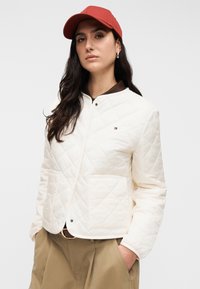 Μπουφάν από μπουκλέ κρέμα με στρογγυλό γιακά, μαύρο κουμπί με πατιλέτα και εμπρόσθια τσέπη· σε συνδυασμό με πουκάμισο σκούρου καφέ και κόκκινο καπέλο.