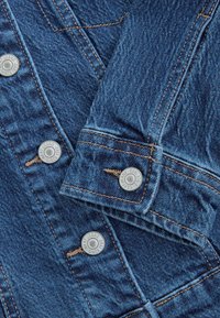 Kék farmerdzseki ujj, fém gombokkal, melyeken a „NEXT JEANS SINCE 1982” dombornyomott, barna varrással, amely látható a szöveten.