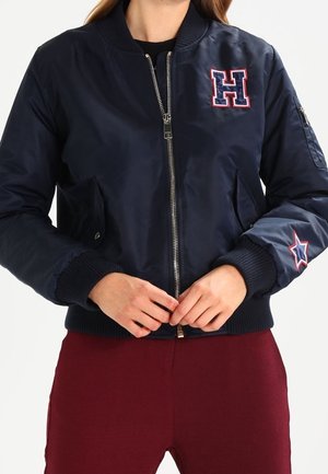 Granatowa kurtka bomber z zamkiem błyskawicznym, ściągaczami na rękawach, naszywką w kształcie litery "H" na piersi, naszywką gwiazdy na rękawie, noszona z bordowymi spodniami.