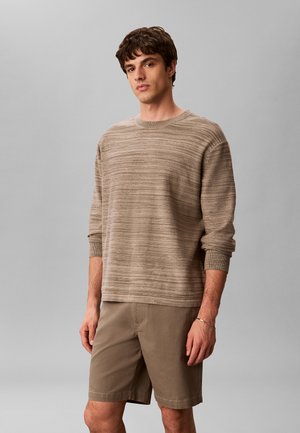 Młody mężczyzna stoi, ubrany w beżowy, pasiasty sweter z długim rękawem oraz pasujące beżowe krótkie spodenki na jednolitym szarym tle.