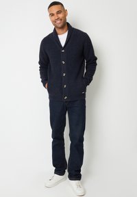 Cardigan lavorato a maglia di colore blu navy con collo a scialle, chiusura frontale con cinque bottoni e trama a costine orizzontali. Abbinato a jeans scuri e sneakers bianche.