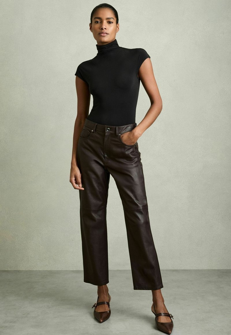 Zwarte, strakke coltruik gecombineerd met hoge, donkere leren broek. Het model draagt bruine, puntige flats, wat een elegante en moderne uitstraling geeft.