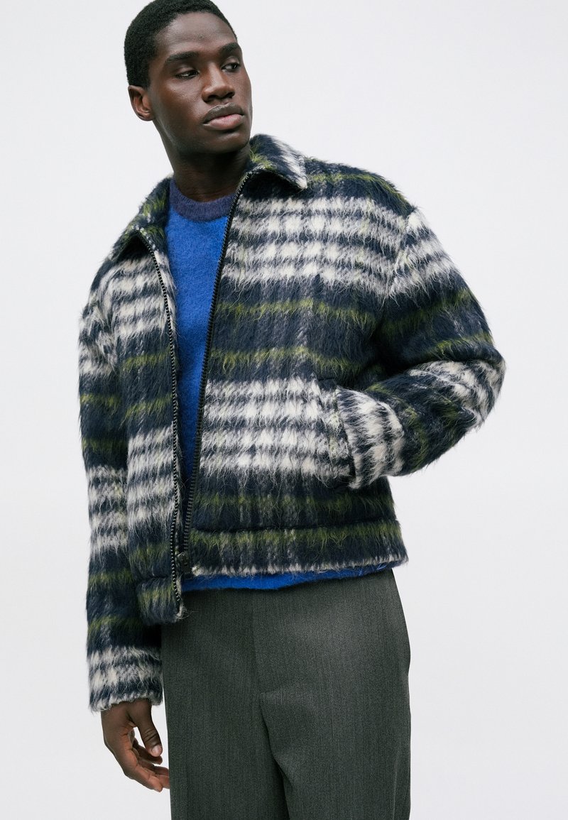 Hombre con una chaqueta de cuadros de felpa y cremallera en azul marino, blanco y verde, un suéter azul y pantalones grises, mirando a su izquierda con una mano en el bolsillo.