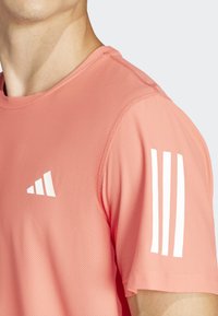 Camiseta atlética de manga corta en color coral, hecha de tejido de malla transpirable. Presenta un logo blanco y tres rayas en la manga derecha.